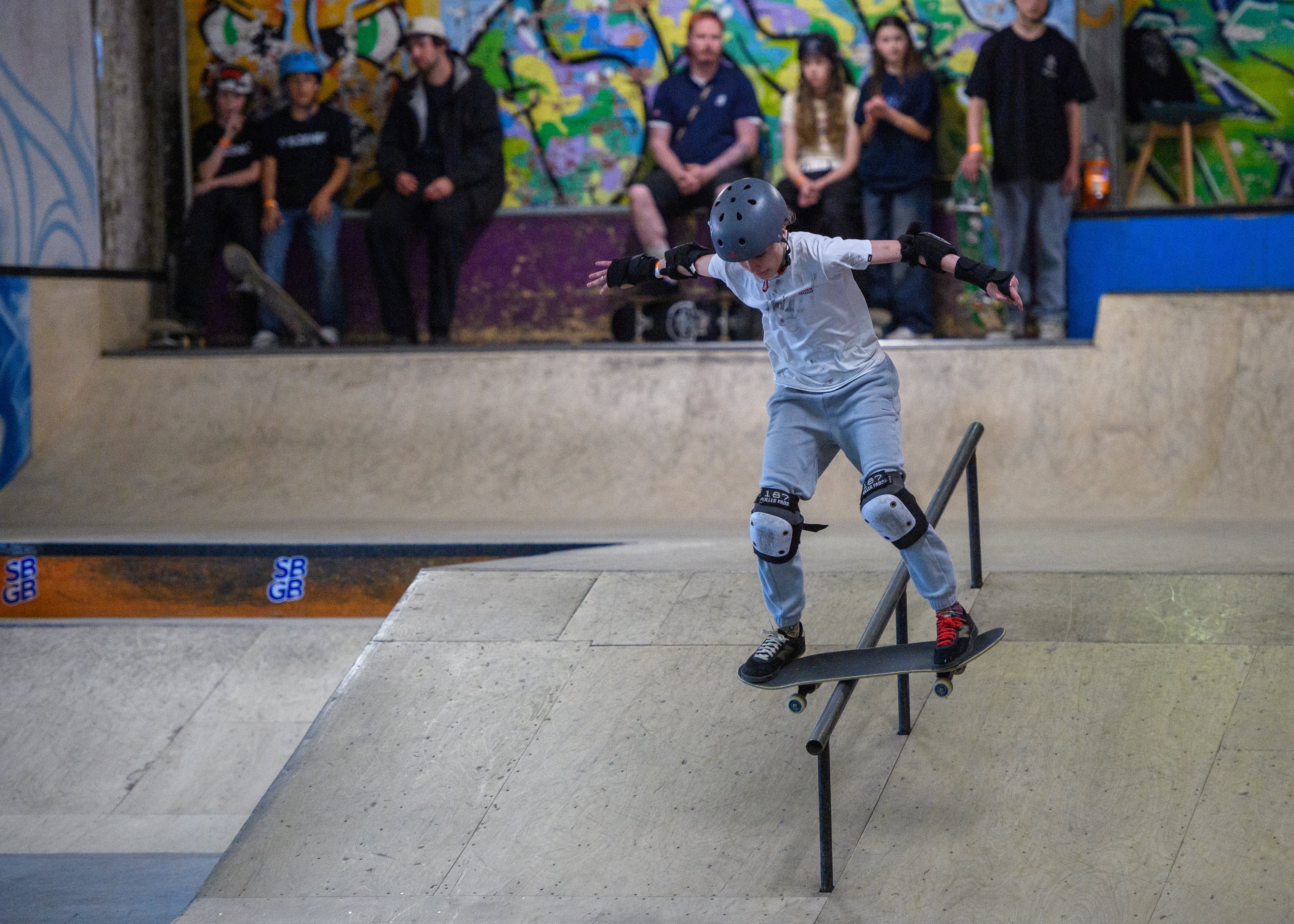 A skateboarder wearing a black helmet, white t-shirt, and light blue jeans performs a trick on a metal rail inside a concrete skatepark, with spectators watching from behind.
